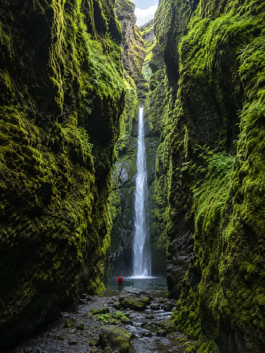 Eine schmale, moosige Schlucht mit einem dünnen Wasserfall am Ende und einem Wanderer als Größenvergleich
