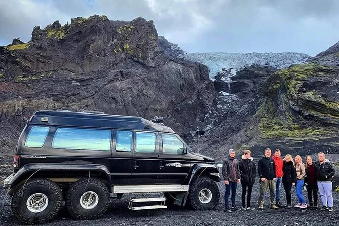 A six-wheel super jeep parked on gravel with a small tour group standing beside it