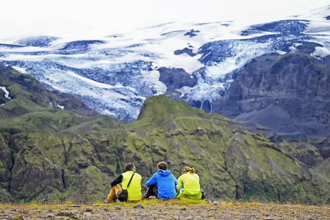 Drei Personen in Outdoorjacken sitzen an einem Hang mit Blick auf einen Gletscher